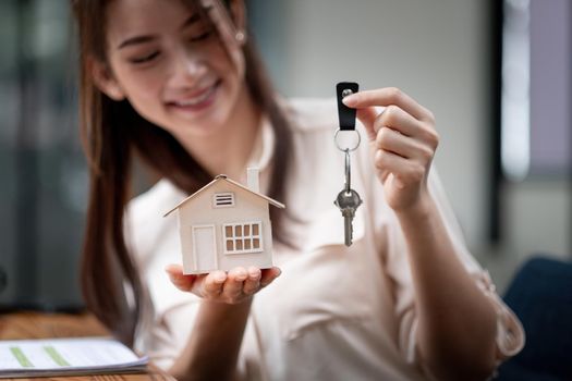 mortgage, real estate and property concept - close up of hands holding house model and home keys at modern office.