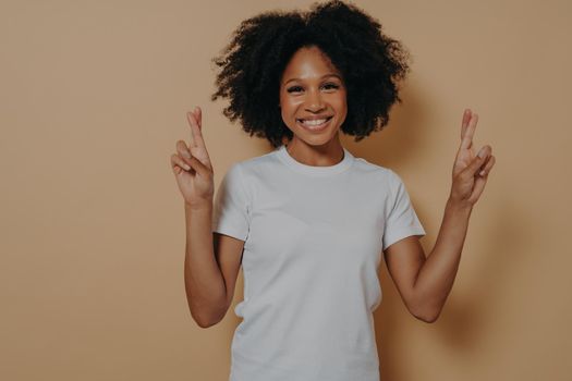 Young hopeful african american female isolated on beige studio background crossing fingers and making wish, showing hope gesture and smiling at camera, asks for good luck. Superstition concept