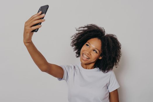 Cheerful young african woman with curly hair taking selfie on her modern mobile phone, looking at camera smiling happily while posing isolated over white studio background, wearing white basic tshirt