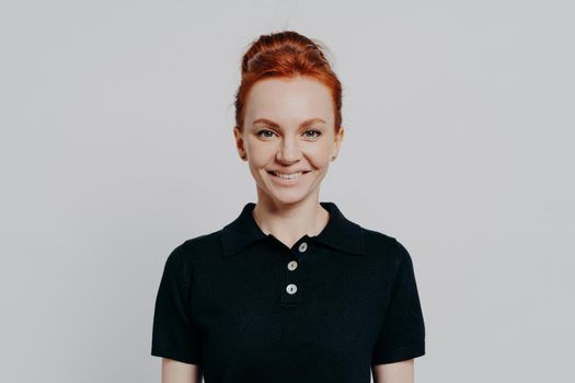 Pretty and positive young red-haired female in black polo t-shirt feeling excited and optimistic while looking at camera, standing isolated over grey background. Happy ginger woman posing in studio