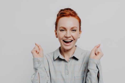 Happy red haired 30s woman wearing casual shirt holding clenched fists and looking at camera with overjoyed emotion, enjoying moment of success and celebrating victory, isolated on grey background