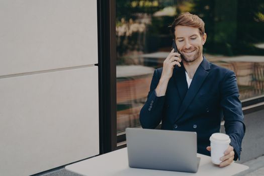 Smiling successful business owner working on laptop remotely and talking with employee on mobile phone, wearing formal suit, sitting in sidewalk cafe and making phone call, holding white coffee cup