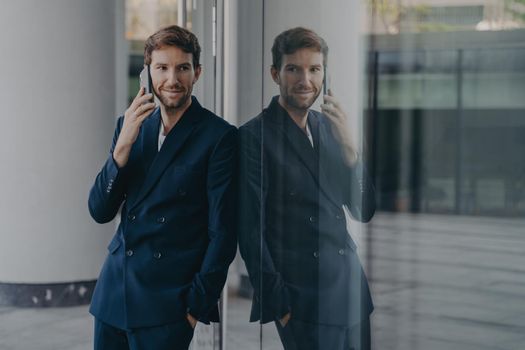 Indoor shot of thoughtful unshaven man employee uses mobile phone for communication arranges meeting with partner poses in modern office building dressed in formal wear. Distant conversation