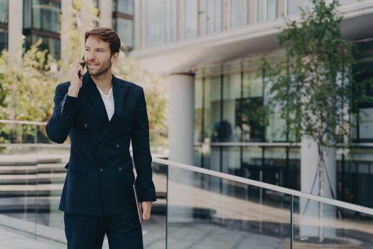 Young confident office worker in formal smart suit walking outside of business center, talking on mobile phone, holding laptop in his hand, hurrying to work on sunny morning