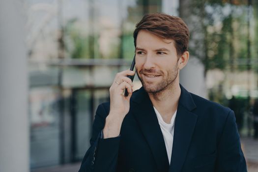 Handsome unshaven male executive worker left business building hears important information via speed mobile connectivity dressed in formal clothes poses against blurred background uses roaming tariffs