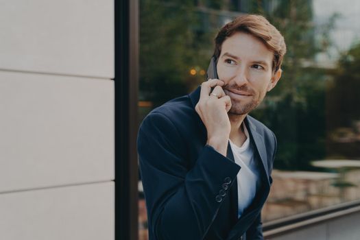 Business communication. Young handsome urban professional businessman listening to customer on cell phone, discussing something on smartphone, standing alone next to building with reflective window