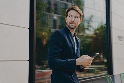 Pensive young businessman in dark suit holding smartphone, thinking about how to reply to partner, looking away while standing outside next to office building, selective focus on man with mobile phone