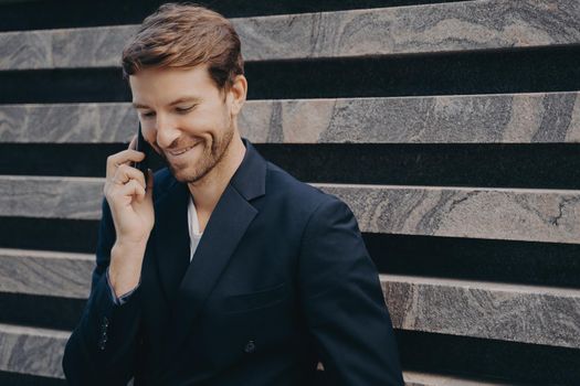 Young attractive confident businessman with neat beard in formal wear speaking on mobile leans against outer wall of city building. Smiling man having conversation on phone while having work break