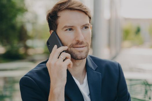 Horizontal shot of male entrepreneur or manager consults client by telephone call discusses future project has self confident expression poses outdoor wears formal clothes has busy working day