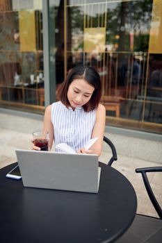 Beautiful confident woman in casual clothing reading book and drinking coffee while sitting in cafe with laptop and notebook 
