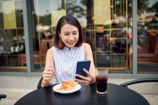 Smiling pretty young woman in casual shirt sitting at table in cafe and checking messenger while using smartphone