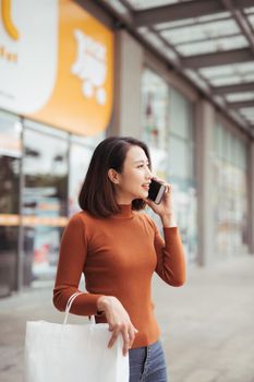 Portrait of happy young Asian woman talking on mobile and carrying paper shopping bags walking outdoor mall