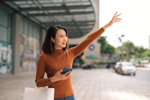 Cheerful young Asian woman with shopping bag catching taxi outside mall