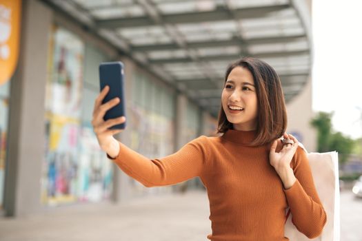 Beautiful young Asian woman holding phone to selfie with shopping bag outdoor shopping mall