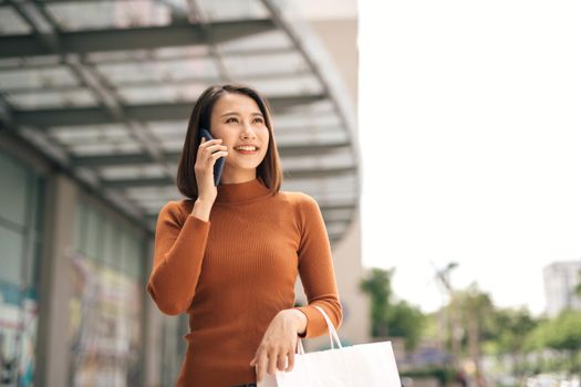 Portrait of happy young Asian woman talking on mobile and carrying paper shopping bags walking outdoor mall