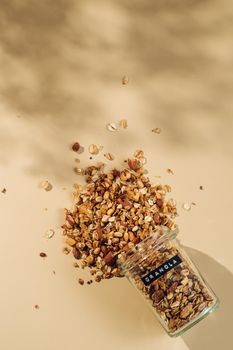 Homemade granola sprinkled of glass jar over pastel beige background. Vertical. Top view or flat lay. Aesthetic oat granola, beautiful shadows, champagne yellow background. Healthy breakfast concept