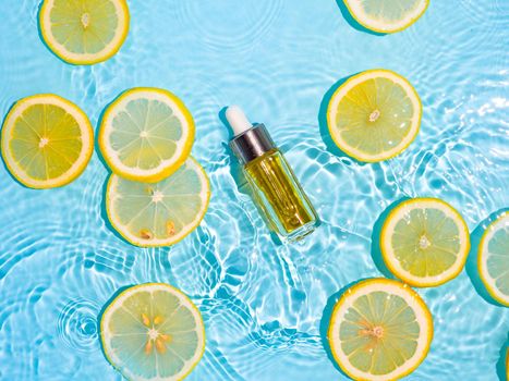Luxury cosmetic oil or serum in glass bottle with pipette in clean transparent water with lemon slices over blue background. Water splashing on water surface in sunlight. Vertical flatlay copy space