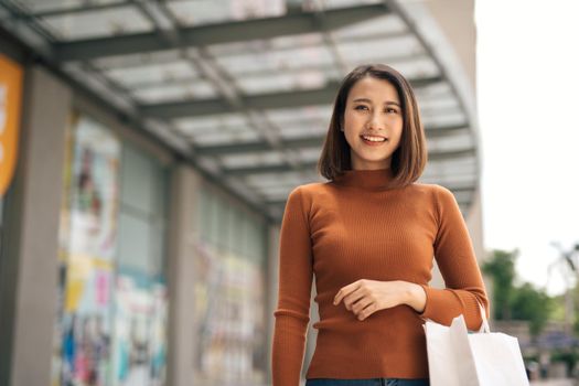 happiness, consumerism, sale and people concept - smiling young Asian woman with shopping bags at outdoor shopping mall