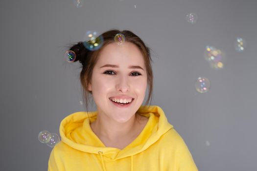 happy brunette woman in yellow hoodie among soap bubbles in bright grey room. happy people. millennial generation. fashionable teenager. trendy colours