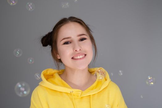 happy brunette woman in yellow hoodie among soap bubbles in bright grey room. happy people. millennial generation. fashionable teenager. trendy colours