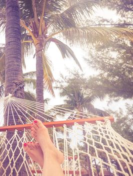 Female legs in a hammock on a background of the sea, palm trees and sunset. Vacation concept with copy space and orange color tone