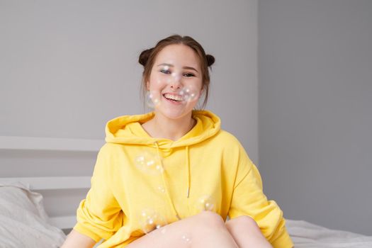 happy brunette woman in yellow hoodie among soap bubbles in bright grey room. happy people. millennial generation. fashionable teenager. trendy colours