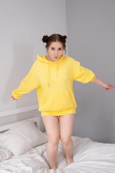 cheerful positive brunette woman in yellow hoodie in white bedroom on bed with white linens. happy people. millennial generation. fashionable teenager.