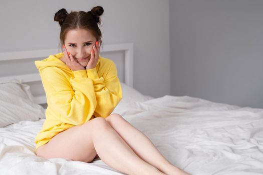 happy cheerful brunette girl in yellow hoodie in white bedroom sitting on bed with white bed linens