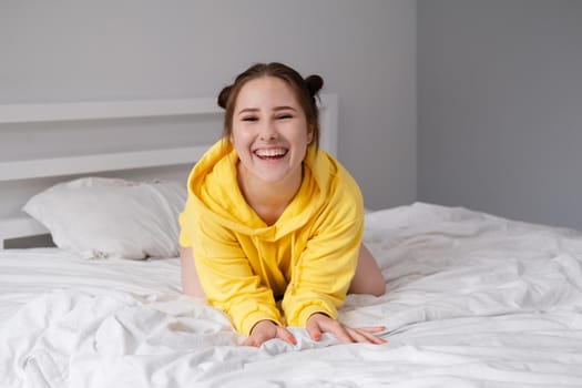 cheerful positive brunette woman in yellow hoodie in white bedroom on bed with white linens. happy people. millennial generation. fashionable teenager.