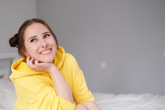 cheerful positive brunette woman in yellow hoodie in white bedroom on bed with white linens. happy people. millennial generation. fashionable teenager.