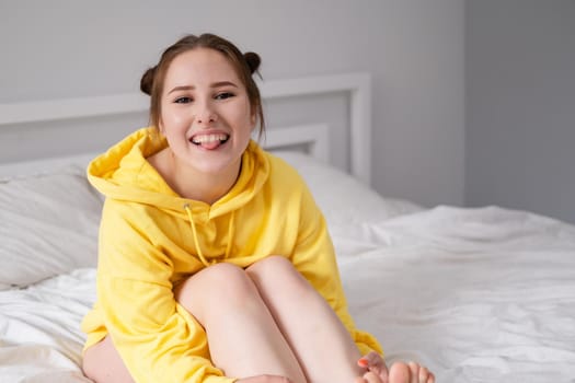 cheerful positive brunette woman in yellow hoodie in white bedroom on bed with white linens. happy people. millennial generation. fashionable teenager.