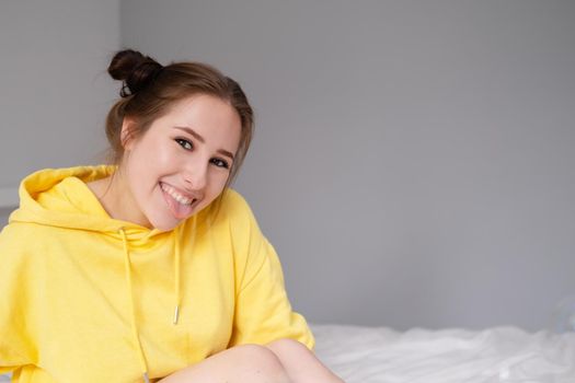 cheerful positive brunette woman in yellow hoodie in white bedroom on bed with white linens. happy people. millennial generation. fashionable teenager.