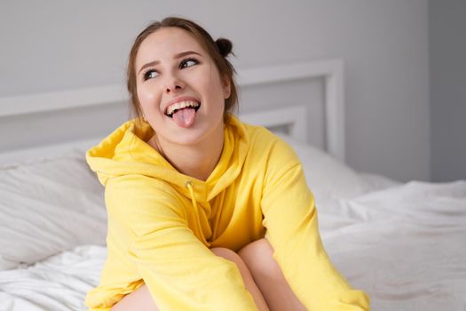 cheerful positive brunette woman in yellow hoodie in white bedroom on bed with white linens. happy people. millennial generation. fashionable teenager.