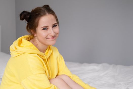 happy cheerful brunette girl in yellow hoodie in white bedroom sitting on bed with white bed linens