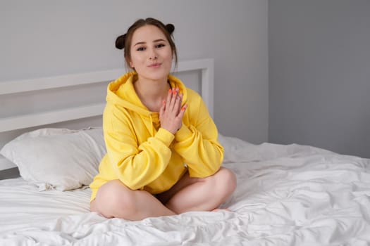 happy cheerful brunette girl in yellow hoodie in white bedroom sitting on bed with white bed linens