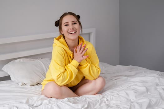 cheerful positive brunette woman in yellow hoodie in white bedroom on bed with white linens. happy people. millennial generation. fashionable teenager.