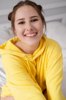 cheerful positive brunette woman in yellow hoodie in white bedroom on bed with white linens. happy people. millennial generation. fashionable teenager.