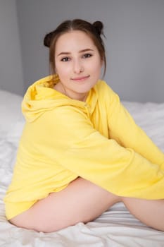 cheerful positive brunette woman in yellow hoodie in white bedroom on bed with white linens. happy people. millennial generation. fashionable teenager.