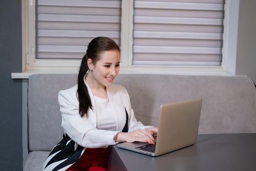 woman sitting by the table on couch in grey room - office and use laptop. big boss. important, confident women. femininity. millennials.