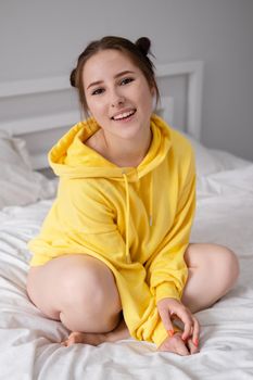 happy cheerful brunette girl in yellow hoodie in white bedroom sitting on bed with white bed linens