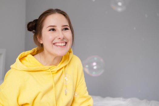 happy brunette woman in yellow hoodie among soap bubbles in bright grey room. happy people. millennial generation. fashionable teenager. trendy colours