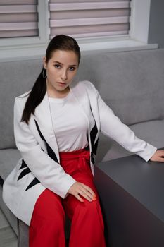 beautiful business woman in red pants, white blouse and Blazer sitting by the table on couch in grey room office.