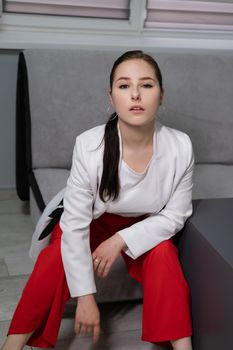 beautiful business woman in red pants, white blouse and Blazer sitting by the table on couch in grey room office.