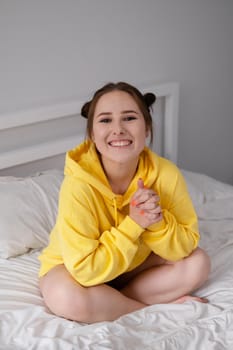 happy cheerful brunette girl in yellow hoodie in white bedroom sitting on bed with white bed linens