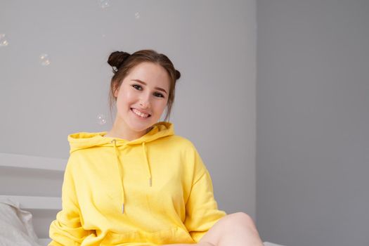 cheerful positive brunette woman in yellow hoodie in white bedroom on bed with white linens. happy people. millennial generation. fashionable teenager.