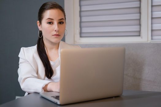 woman sitting by the table on couch in grey room - office and use laptop. big boss. important, confident women. femininity. millennials.