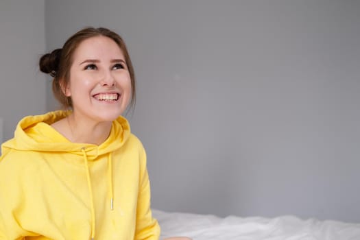 cheerful positive brunette woman in yellow hoodie in white bedroom on bed with white linens. happy people. millennial generation. fashionable teenager.