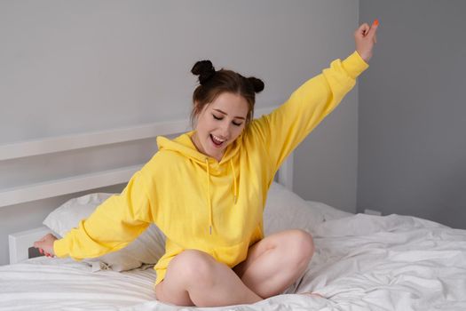 happy cheerful brunette girl in yellow hoodie in white bedroom sitting on bed with white bed linens