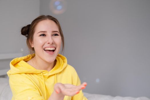 happy brunette woman in yellow hoodie among soap bubbles in bright grey room. happy people. millennial generation. fashionable teenager. trendy colours