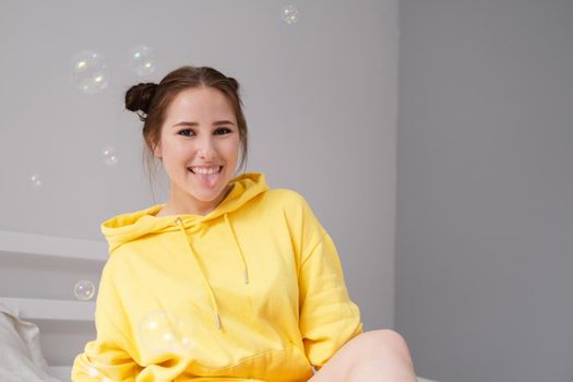 cheerful positive brunette woman in yellow hoodie in white bedroom on bed with white linens. happy people. millennial generation. fashionable teenager.